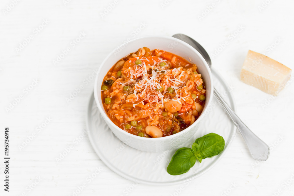 Bowl of Minestrone, a traditional Italian vegetable soup made of carrots, peas, beans, celery, tomatoes, garlic and onions with grated parmesan cheese and basil on white kitchen table Bowl of Minestrone, a traditional Italian vegetable soup made of carrots, peas, beans, celery, tomatoes, garlic and onions with grated parmesan cheese and basil on white kitchen table