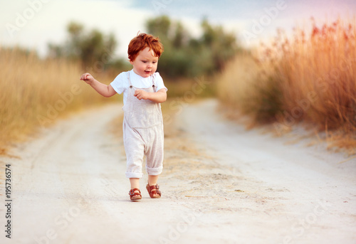 adorable redhead toddler baby boy in jumpsuit running along rural summer road in sunburned field