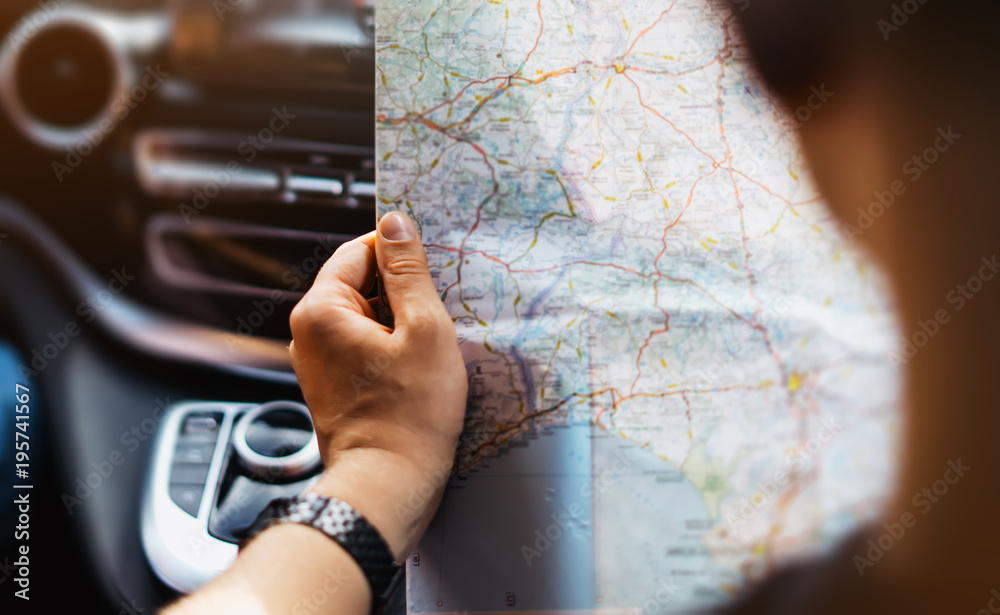 Obraz premium Hipster man holding in male hands and looking on navigation map in auto, tourist traveler hiker driving on background panoramic view way road, trip in transportation, person sitting in auto