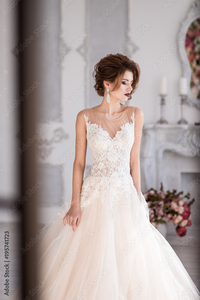 Morning of the bride. A beautiful bride is standing in a lace wedding dress. Wedding hairstyle and makeup. Photo through the window. Morning of the bride. A beautiful bride is standing in a lace wedding dress. Wedding hairstyle and makeup. Photo through the window.