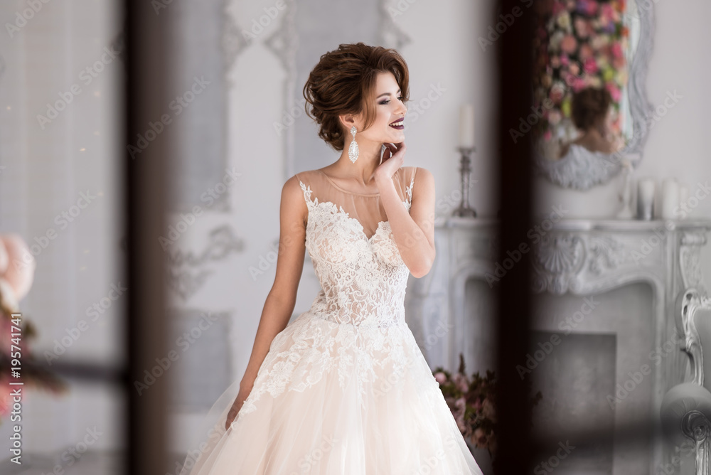 Morning of the bride. A beautiful bride is standing in a lace wedding dress. Wedding hairstyle and makeup. Hand at the face. Photo through the window. Morning of the bride. A beautiful bride is standing in a lace wedding dress. Wedding hairstyle and makeup. Hand at the face. Photo through the window.