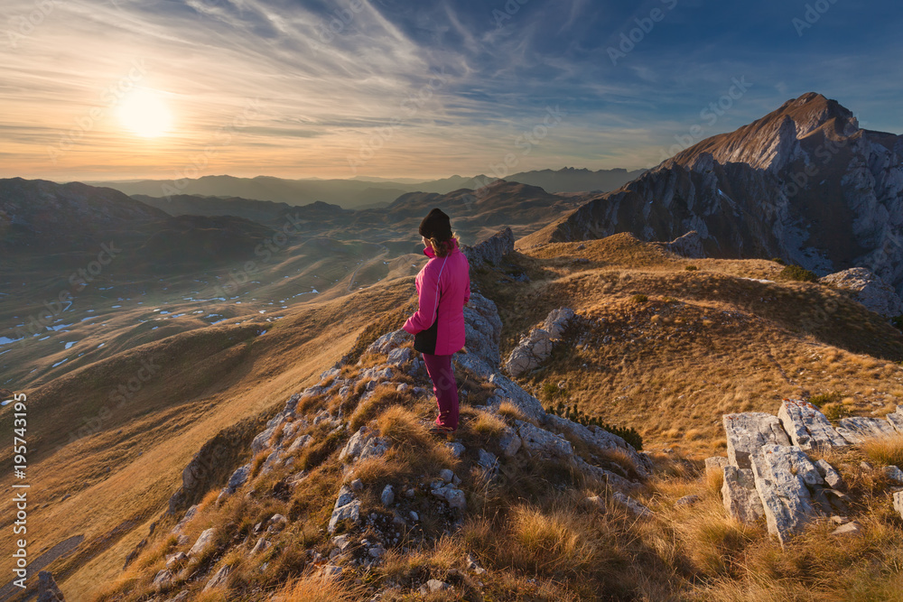 Woman hiker standing on the mountain cliff towards the setting sun