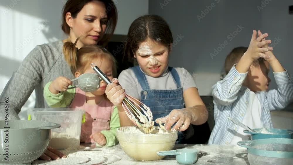 A beautiful mother with three cheerful children of different ages kneads the dough. The elder daughter removes the dough from the corolla, the younger sister helps knead the dough A beautiful mother with three cheerful children of different ages kneads the dough. The elder daughter removes the dough from the corolla, the younger sister helps knead the dough