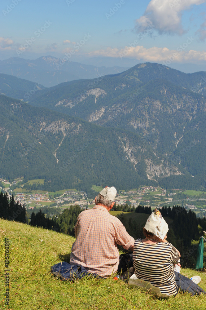 Older couple sitting on the grass looking at the valley and the mountains.