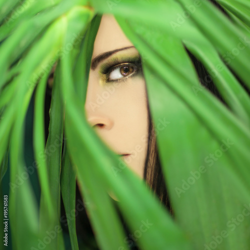 beautiful woman looking through jungle palm leaves, perfect skin and perfect make up, studio portrait in green