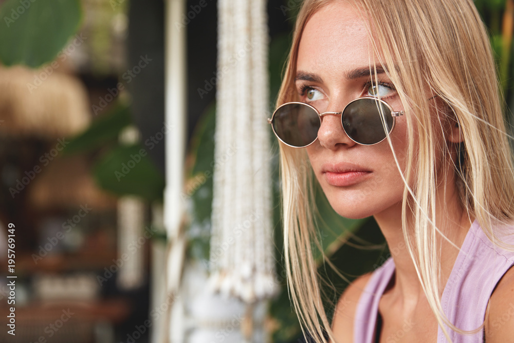 Sideways portrait of thoughtful stylish hipster girl in trendy sunglasses, looks pensively away, contemplates about future plans. Beautiful blonde woman being deep in thoughts. People, style concept