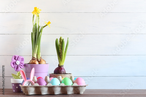 Fototapeta Naklejka Na Ścianę i Meble -  Easter scene with colorful eggs, a bunny clock, and potted flowers against a white plank background. Concept of: Springtime arrangement. Easter still life.