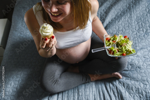 Pregnant woman holds green salad with tomatos in one hand and eating tasty sweet cupcake photo