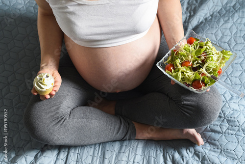 Pregnant woman holds green salad with tomatos and tasty sweet cupcake in her hands photo