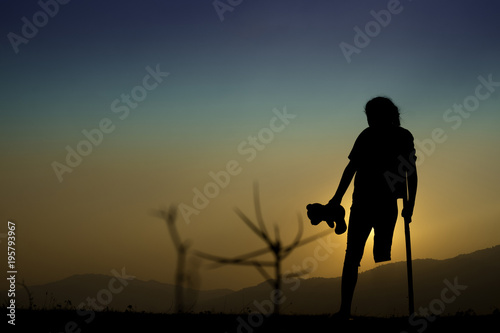 Child disabled with crutches and teddy bear in hand on hill at sunset time. Concept help.