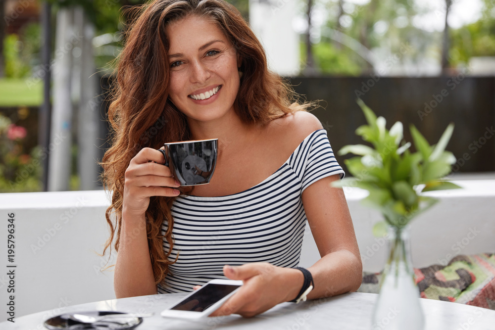 Lovely happy woman sends text messages on smart phone, recreats against coffee shop interior with hot beverage, being in high spirit as browses websites and connected to high speed internet.