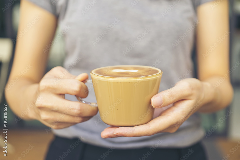 Young entrepreneur making hot coffee and serving to customer. Woman with gray shirt holding a brown cup of coffee which made from ceramic with decoration on top of mug. Young entrepreneur making hot coffee and serving to customer. Woman with gray shirt holding a brown cup of coffee which made from ceramic with decoration on top of mug.