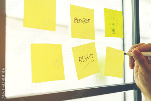 Hand holding pen and writing on sticky note papers which are on mirror wall. Wording Budget, Right and drawing lamps are on note paper and empty. Brainstorming teamwork to write financial keyword photo