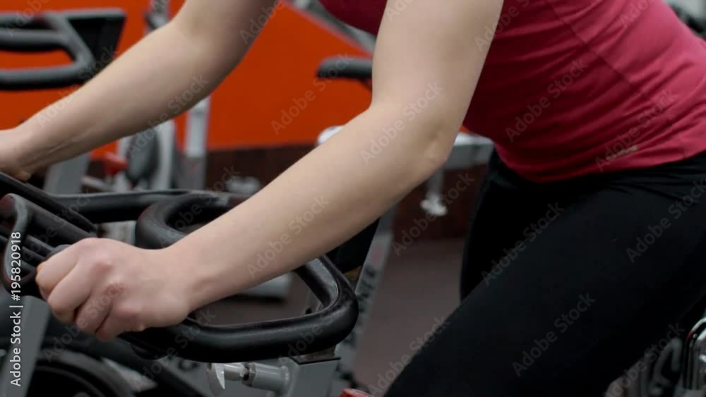 Young woman is having a training on stationary bicycle in modern gym. Lady in red t-shirt and black sport trousers is working on her gluteus and harmstrings and holsing the handlebar of the machine by Young woman is having a training on stationary bicycle in modern gym. Lady in red t-shirt and black sport trousers is working on her gluteus and harmstrings and holsing the handlebar of the machine by
