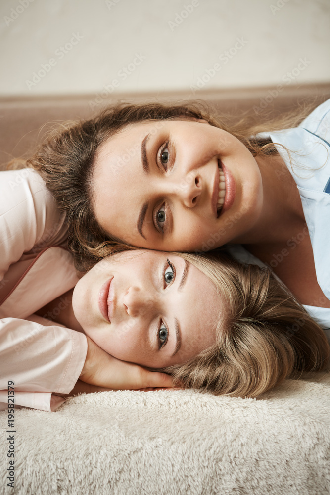 Two girls with close relationship, feeling like sisters. Vertical shot of good-looking women lying on sofa and smiling broadly at camera. Charming curly-haired friend lying on head of her bestie