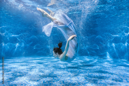 Woman in a white dress dives under the water in the pool.
