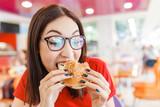Happy healthy woman sitting in indoors food court and eating an delicious hamburger, modern meal concept
