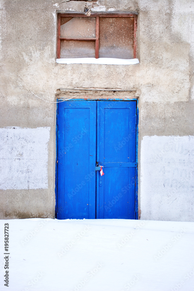 Closed blue painted door with window, gray plaster on wall, grunge background, winter snowy street, vertical Closed blue painted door with window, gray plaster on wall, grunge background, winter snowy street, vertical