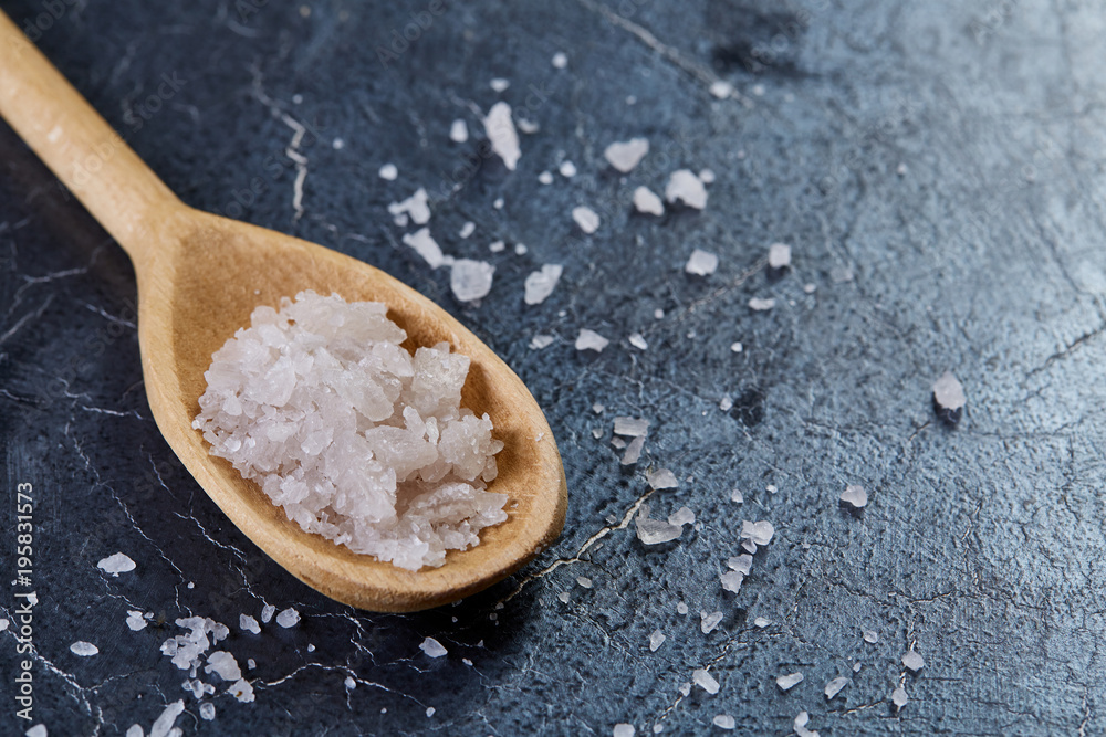 Crystal sea salt in a wooden spoon on dark marble background, top view, close-up, selective focus.
