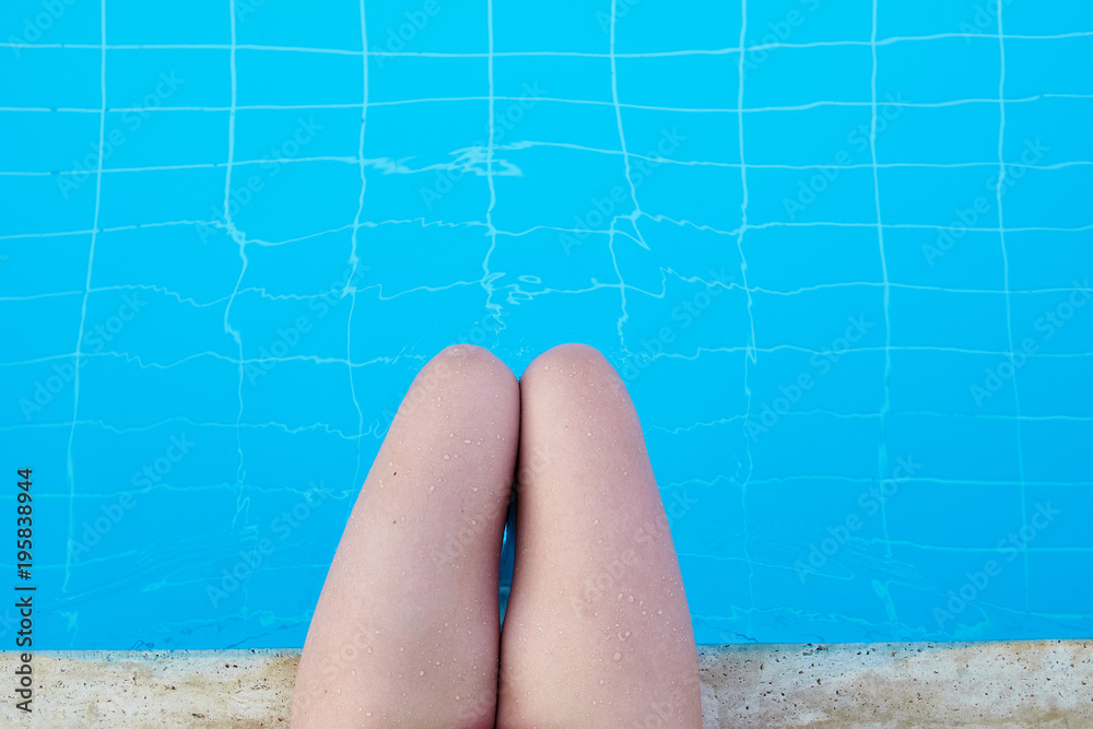 The girl splashes her legs in the pool with clear blue water. A woman is resting by the pool on a hot summer day. Shapely female legs. Women's legs and feet in water.