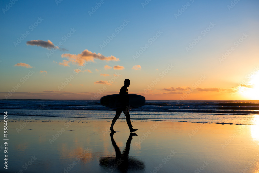 Sunset surfing. Silhouette of man surfer walking with a surf board in his hands across the ocean shore.