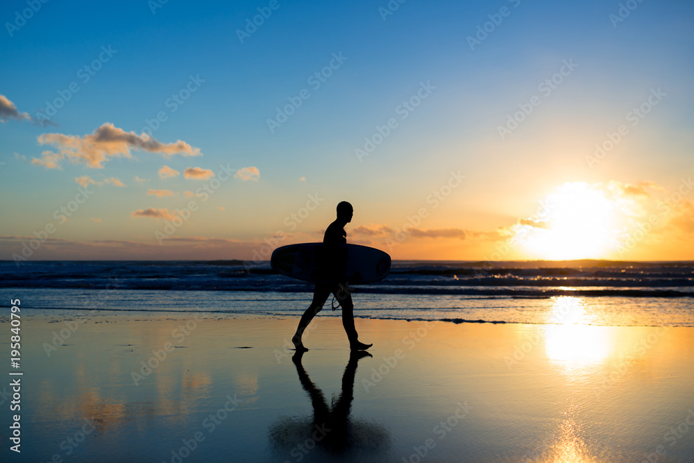 Sunset surfing. Silhouette of man surfer walking with a surf board in his hands across the ocean shore.
