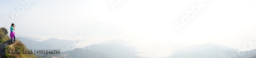 Panorama of woman holding camera and standing on the mountain top looking at the view of beautiful mountains with cloudy sky at Doi Pha Tang, Chiangrai, Thailand