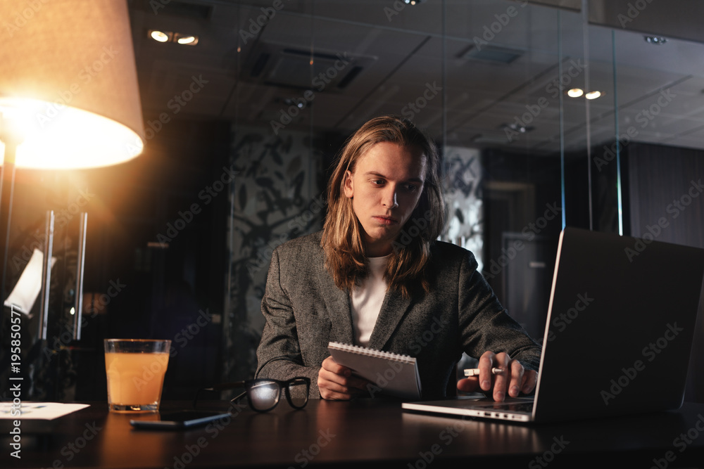 Tired young man with long hair working at night loft office.Coworker work in modern coworking studio. Businessman uses contemporary laptop Tired young man with long hair working at night loft office.Coworker work in modern coworking studio. Businessman uses contemporary laptop