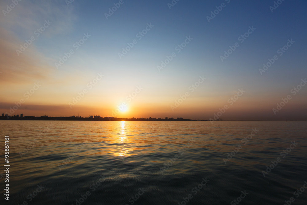 Beautiful sea landscape. Bright warm sunset in the sea near the coastline with the city's silhouettes. Sun hides behind the house. Colors of the sky are reflected in the water