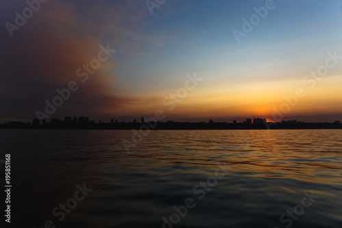Beautiful sea landscape. Bright warm sunset in the sea near the coastline with the city's silhouettes. Sun hides behind the house. Colors of the sky are reflected in the water © ViDi Studio