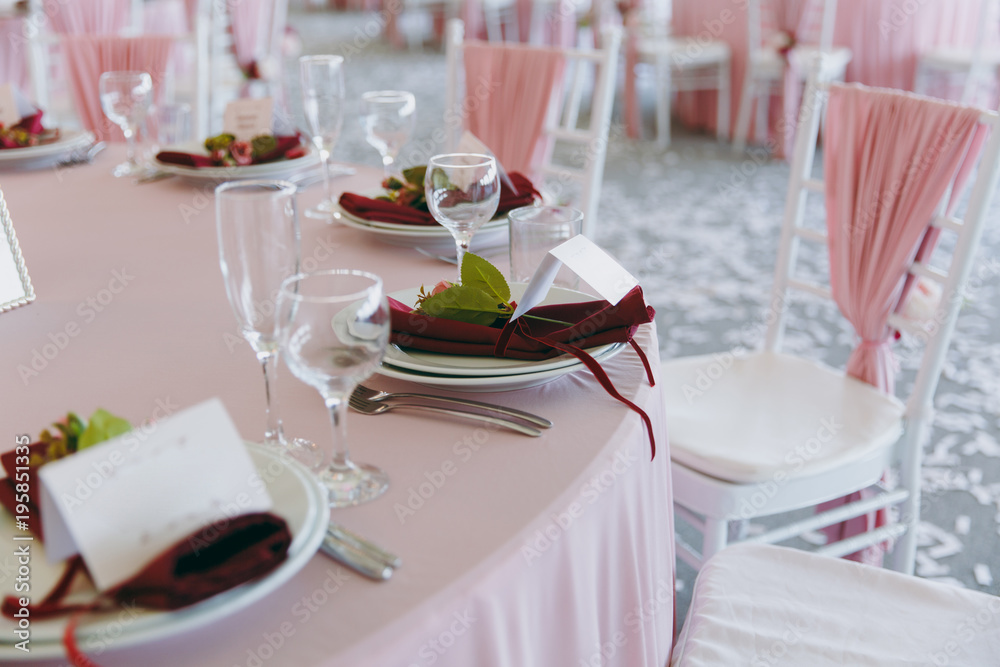 Beautiful decoration of the wedding banquet under the awning in pink, burgundy and white tones. Table setting in a hall with plates, cutlery, glasses, napkins and cards, decorated floral compositions