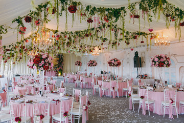 Beautiful decoration of the wedding banquet under the awning in pink, burgundy and white tones. Hall with dining tables and chairs decorated with thin cloth, bouquets, flower garlands and confetti
