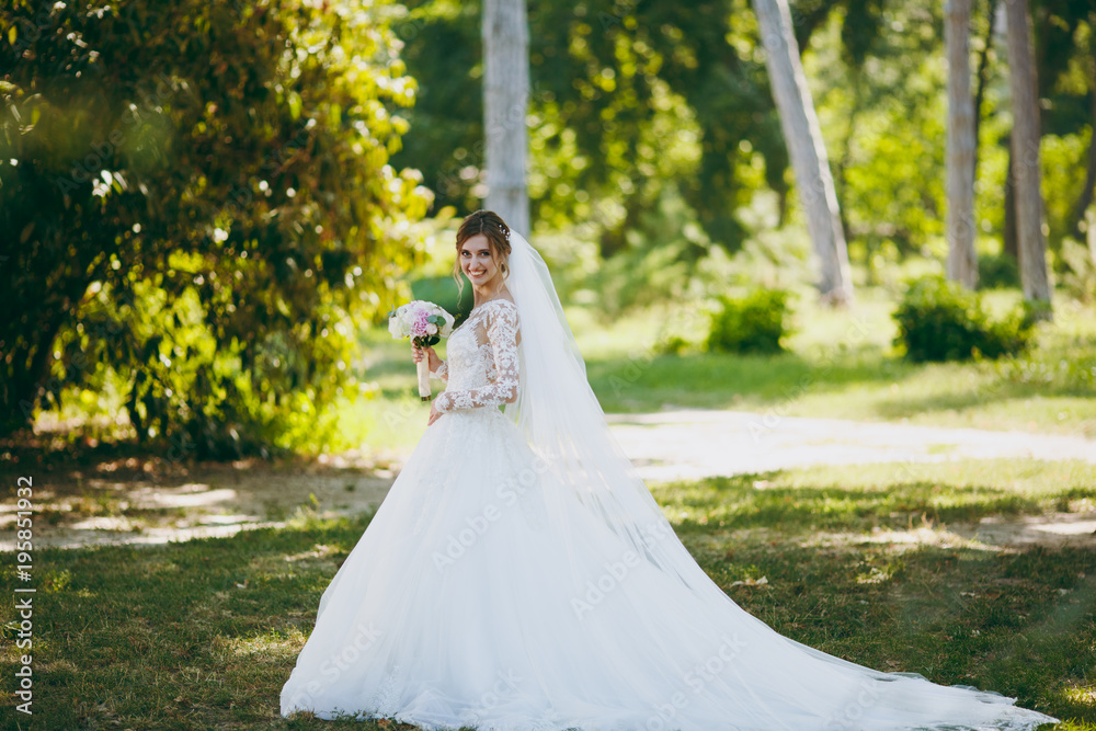 Beautiful wedding photosession. The bride in a white lace dress with a long plume, hairpin in hair, veil and bouquet of white and pink flowers in a large green garden on weathery sunny day Beautiful wedding photosession. The bride in a white lace dress with a long plume, hairpin in hair, veil and bouquet of white and pink flowers in a large green garden on weathery sunny day