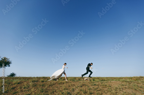 Beautiful wedding photosession. Young charming bride in white lace dress with long plume runs for her fiancé in a black suit on walk around the big green field against blue sky background