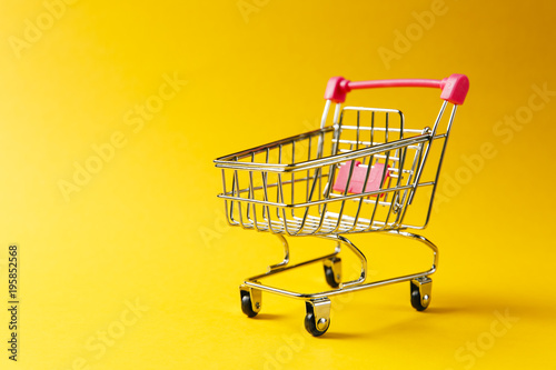 Close up of supermarket grocery push cart for shopping with black wheels and red plastic elements on handle isolated on yellow background. Concept of shopping. Copy space for advertisement