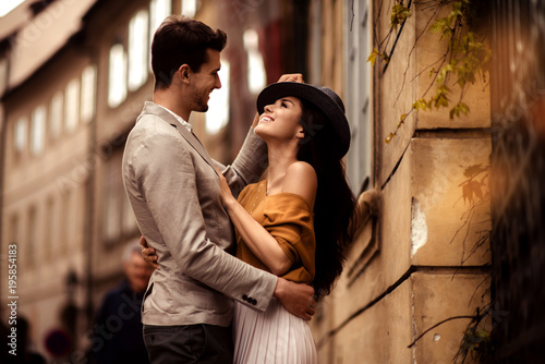 Passionate gorgeous young couple embrace each other while walk across ancient city. Cheerful elegant cute female model wears stylish hat, looks with cheerful expression in her boyfriend s eyes.