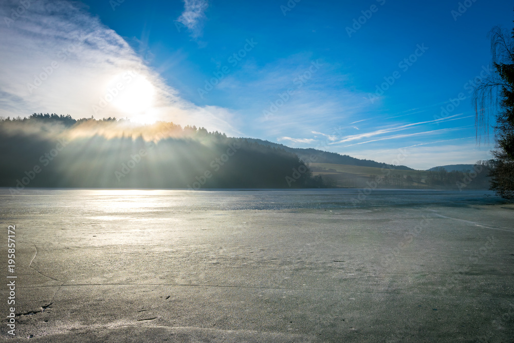 Sonnenstrahlen der Sonne welche auf den gefrorenen See im Bayerischen Wald treffen mit Wälder und Berge im Hintergrund und etwas Schnee auf dem Eis