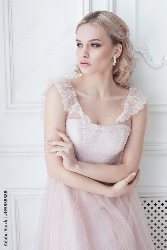Caucasian blonde attractive women in bright white grey interior in light rose gown dress posing on wooden floor Caucasian blonde attractive women in bright white grey interior in light rose gown dress posing on wooden floor