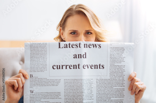 Latest news. Cute pretty young woman hiding behind the page while holding a big newspaper and showing the title of its interesting important article