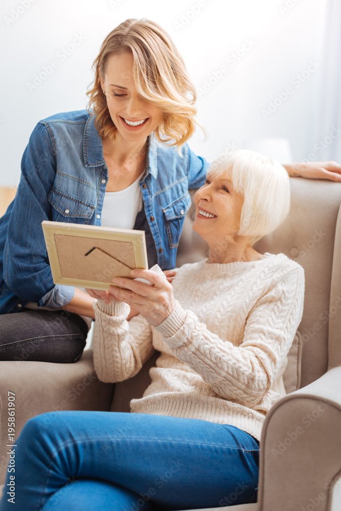 Cute moment. Polite kind attentive young woman smiling and looking at the nice old photo while having a good time with her positive aged grandmother Cute moment. Polite kind attentive young woman smiling and looking at the nice old photo while having a good time with her positive aged grandmother