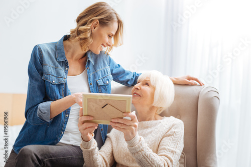 Sharing memories. Kind calm senior woman feeling glad while looking at her attentive loving granddaughter and showing important memorable photos to her