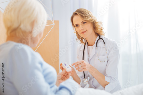 Calm doctor. Experienced clever reliable doctor sitting in front of her ill senior patient and looking friendly while pointing to a convenient tiny pillbox