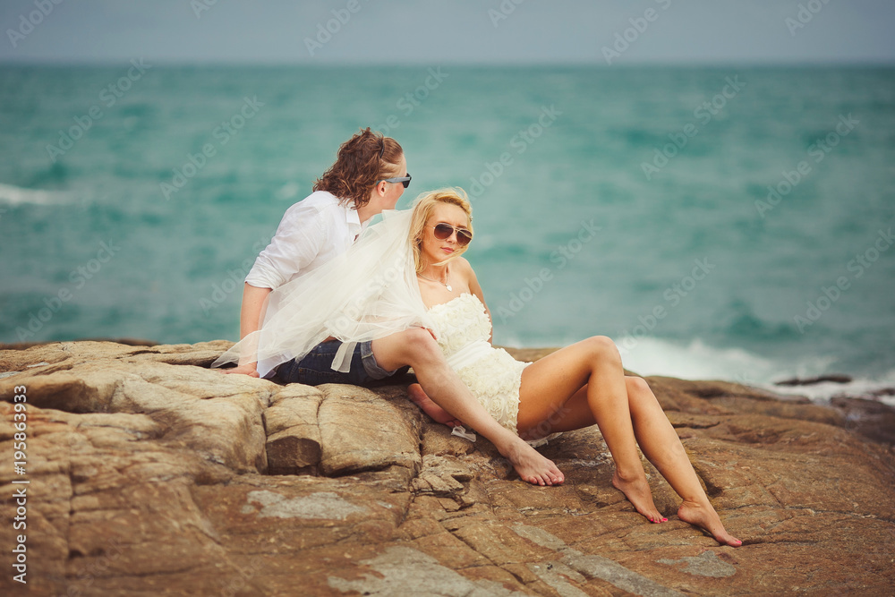 bride and groom sitting on rocky seashore. the newlywed couple is resting in Thailand.