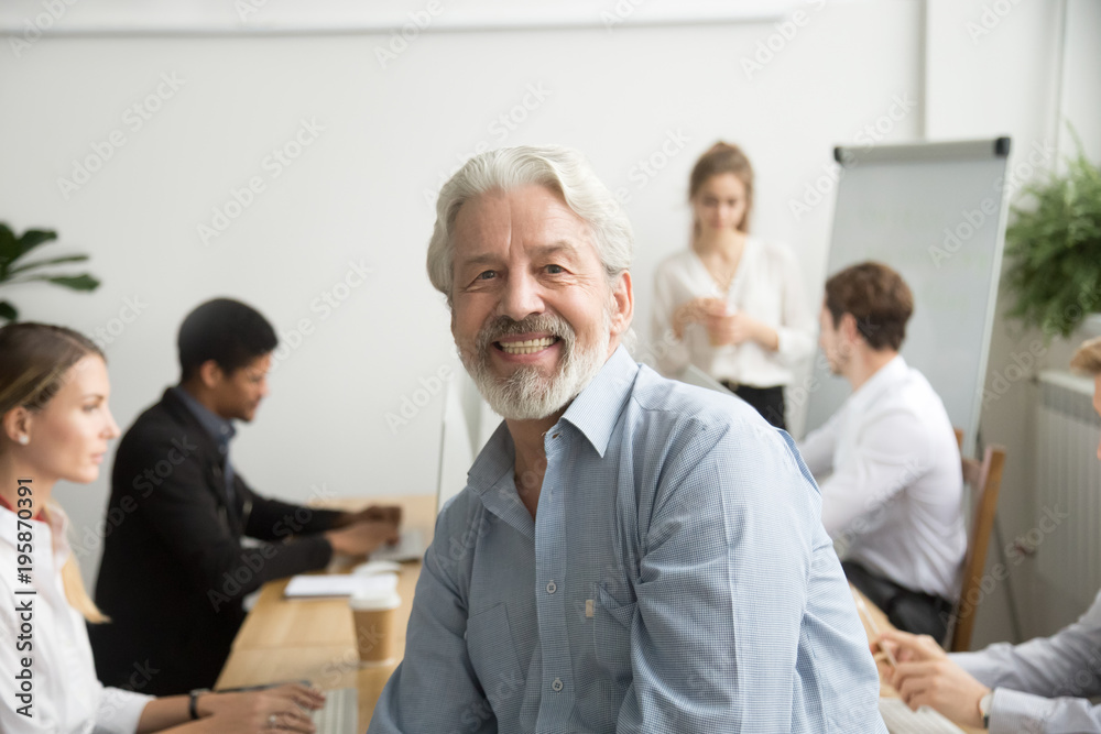 Smiling male senior team leader, aged teacher looking at camera with office people at background, happy old gray-haired company boss, experienced mentor or executive professional head shot portrait Smiling male senior team leader, aged teacher looking at camera with office people at background, happy old gray-haired company boss, experienced mentor or executive professional head shot portrait