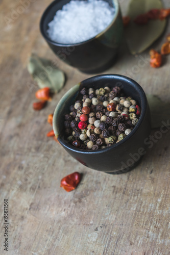 Close up view of spices, coarse salt and colorful allspice pepper in ceramic bowls, bay leaves and dried chilly peppers on old rustic wooden background. Vintage toned. Spices background