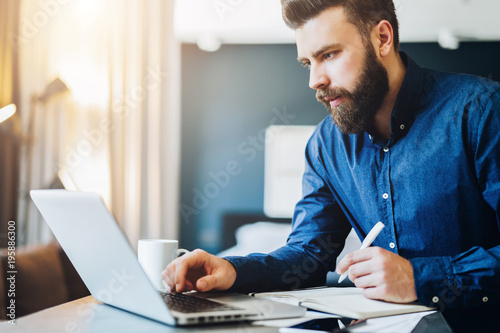 Young bearded businessman works on computer, takes notes in notebook, analyzes information, makes business plan. Freelancer, entrepreneur works at home. Online marketing, education for adults.