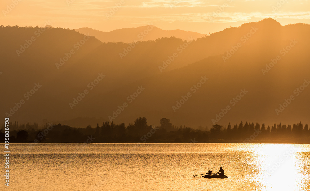 Silhouette scene of fisherman sailing to find fish on small boat in lake with background of big mountain ranges and forest.