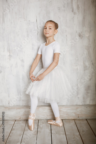 A little adorable young ballerina in white dress and point shoes on woody floor and gray studio background posing on camera