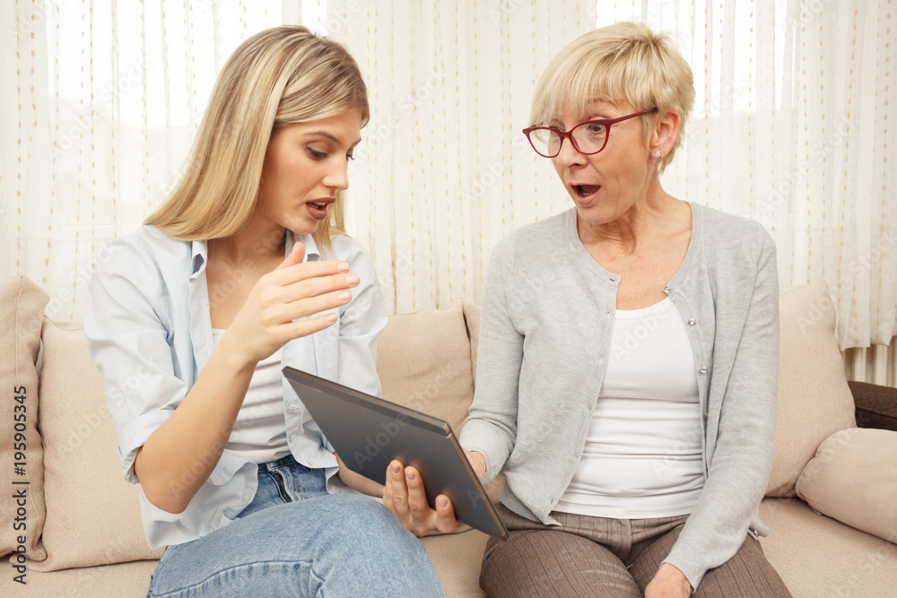 Senior mother is surprised while her daughter teaches her to use a tablet. Happy family moments at home.