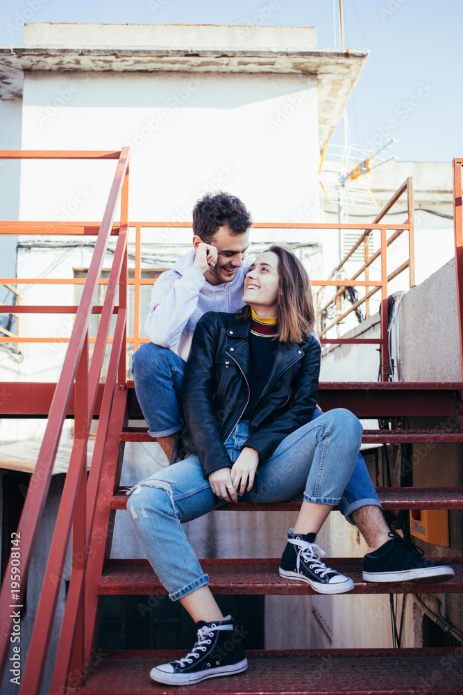 Happy smiling and laughing young couple in trendy fashionable modern clothes sit on red fire escape stairs, pose for model photoshoot on rooftop. Cool and hip teenagers or millennials influencers Happy smiling and laughing young couple in trendy fashionable modern clothes sit on red fire escape stairs, pose for model photoshoot on rooftop. Cool and hip teenagers or millennials influencers